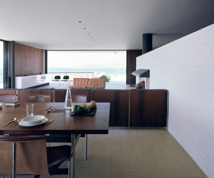 Inside a modern, open-plan 1960s apartment, with a dining area at the front, a lounge area with sofa and fireplace behind it, and a large floor-to-ceiling window overlooking the Mediterranean coast 